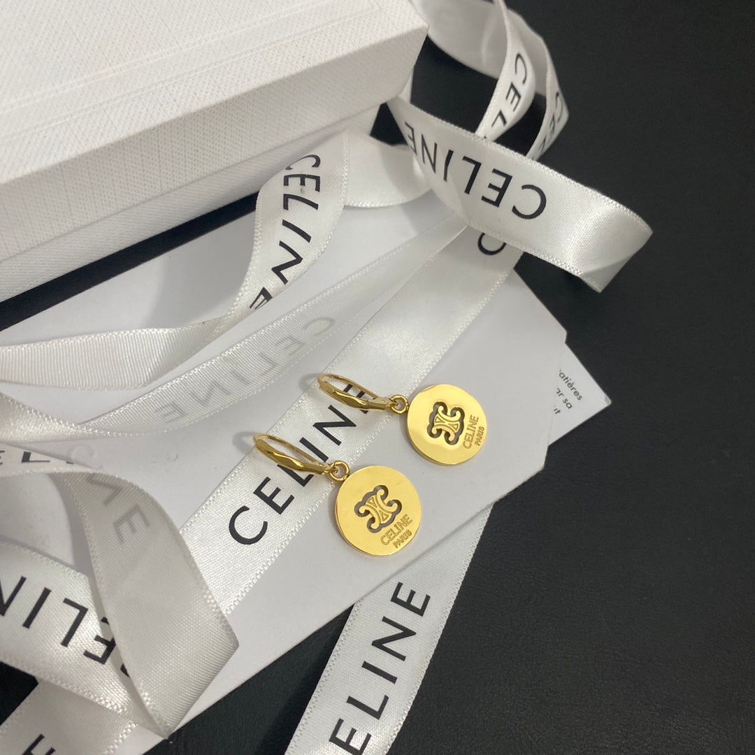 A pair of gold Celine dangle earrings with circular charms featuring the interlocking CC logo, displayed on white Celine branded ribbons and packaging against a dark background.