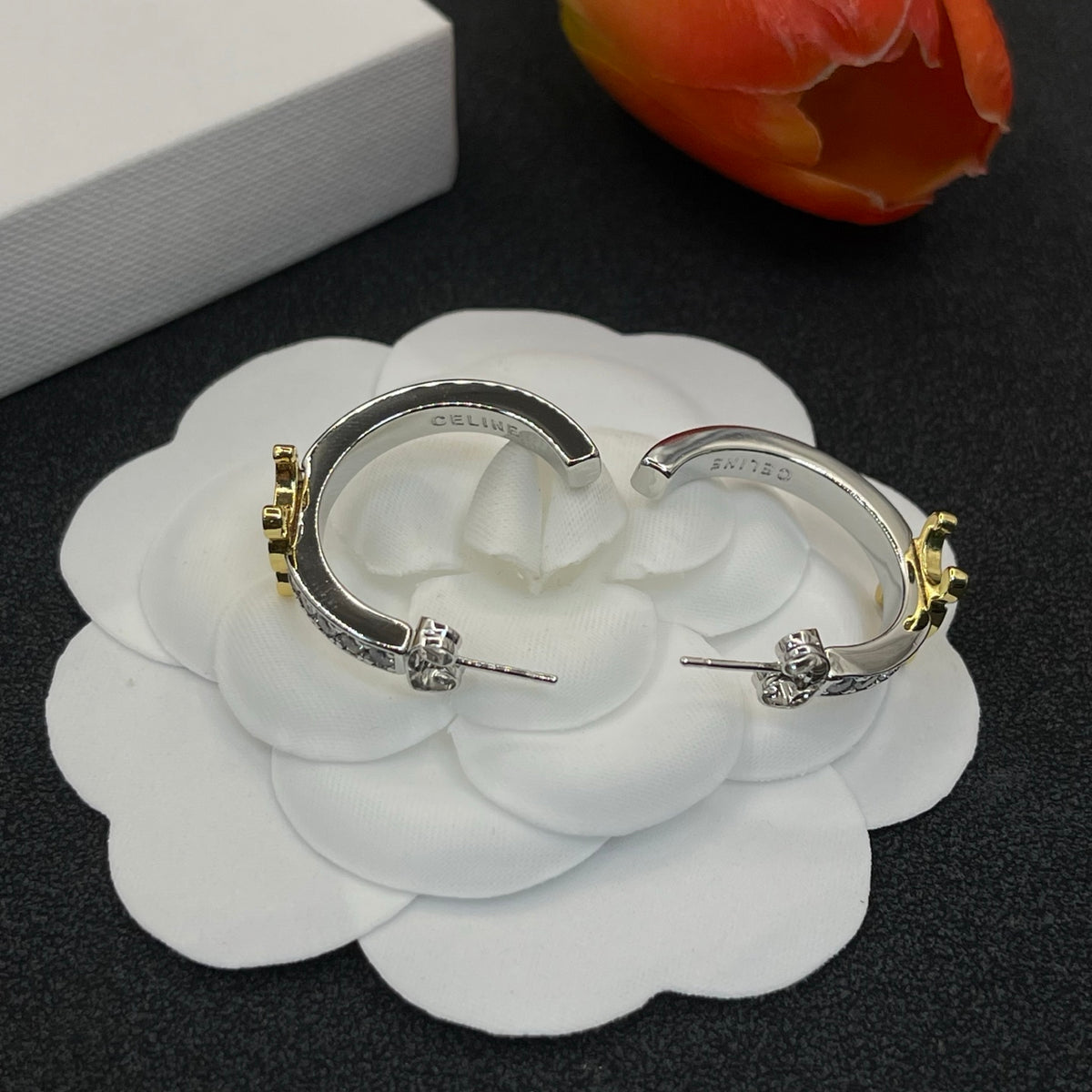 A pair of silver-toned huggie hoop earrings with gold-toned floral accents and small clear stones, displayed on a white fabric flower against a dark background.