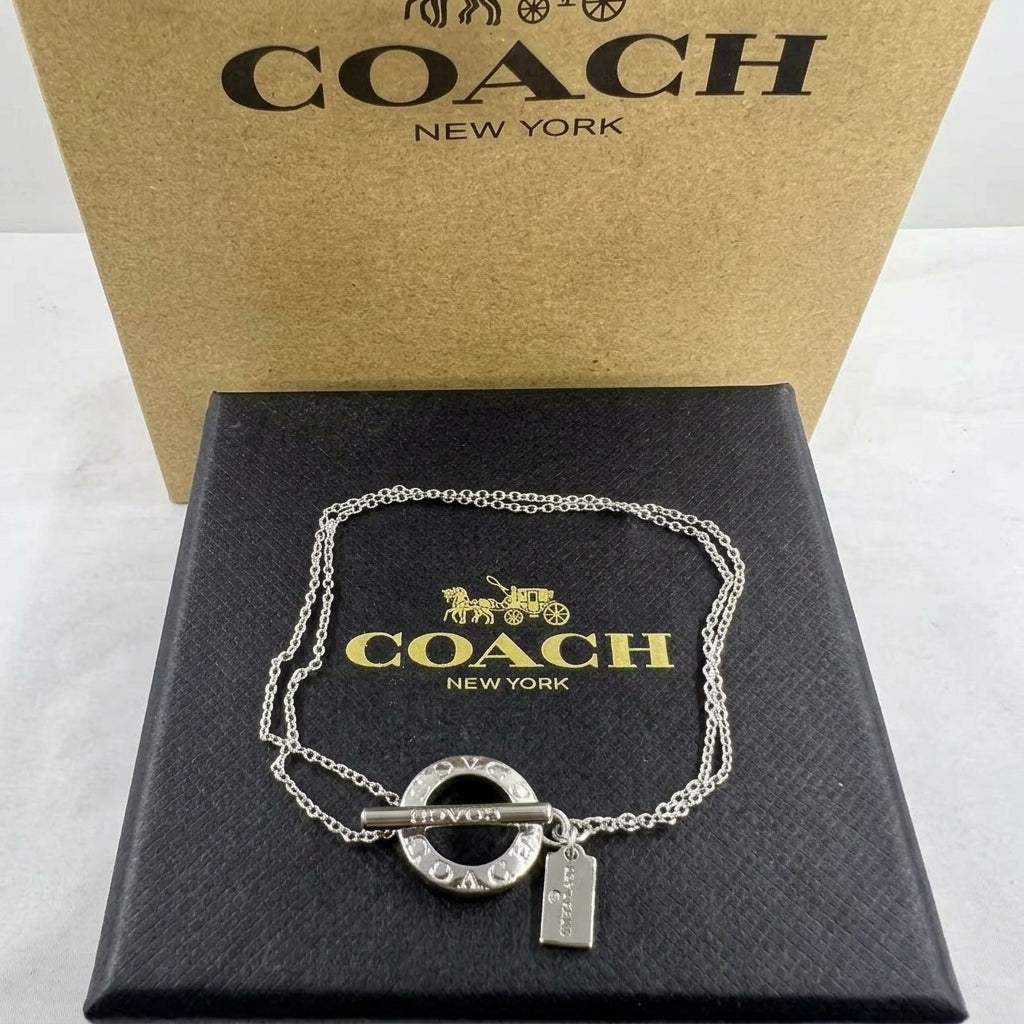 A silver-tone Coach chain necklace with a circular ring, bar toggle clasp, and a small hanging tag, displayed on a black Coach box with a tan Coach box behind it.