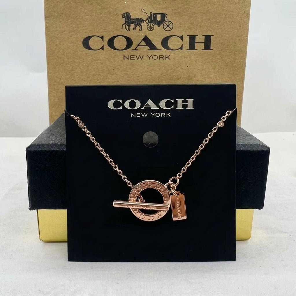 A Coach New York necklace in rose gold, featuring a circular interlocking key and padlock pendant, displayed on a black branded card in front of a brown Coach box.