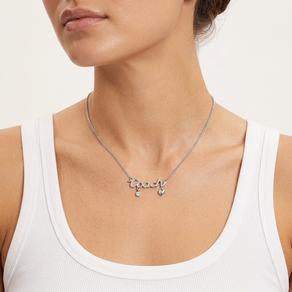A close-up of a model wearing a silver script nameplate necklace that reads 'Coach' with small dangling charms, paired with a white ribbed tank top against a light neutral background.
