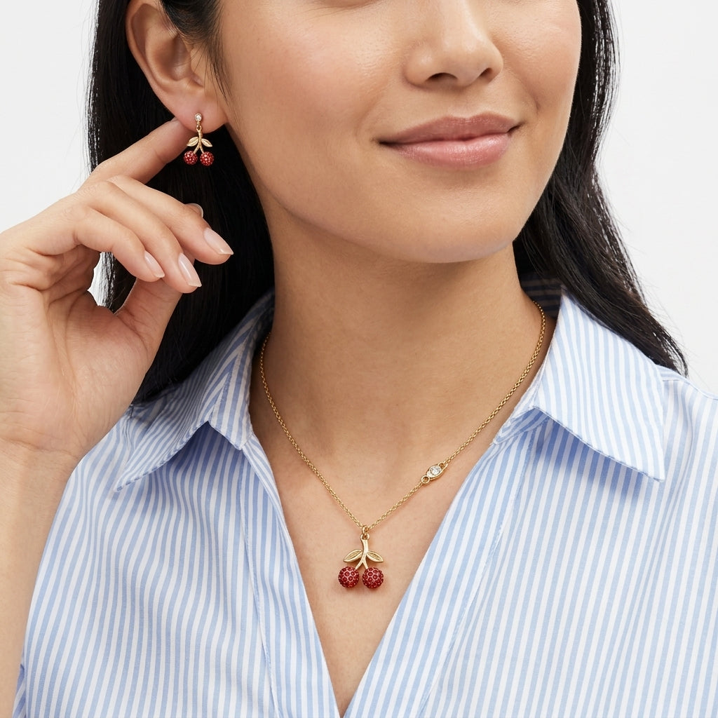 A close-up lifestyle shot of a woman wearing a gold necklace with a red cherry pendant and matching red cherry drop earrings, paired with a blue and white striped shirt.