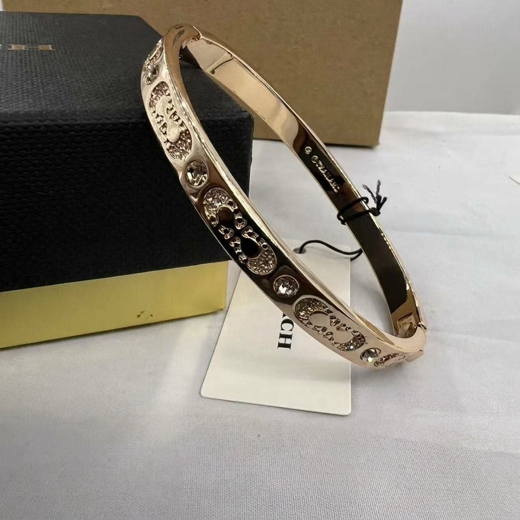 A rose gold bangle bracelet with 'Chanel' lettering and crystal embellishments, displayed on a white surface next to its black and gold Chanel box.