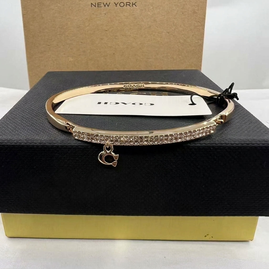 A Coach bangle bracelet in rose gold with a row of clear crystals across the front and a dangling Coach logo charm, displayed on a black presentation box with a Coach tag attached.
