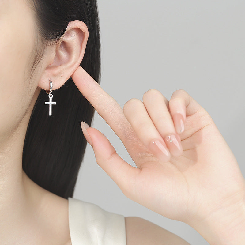 Silver cross earring being worn and held by a hand against a neutral background