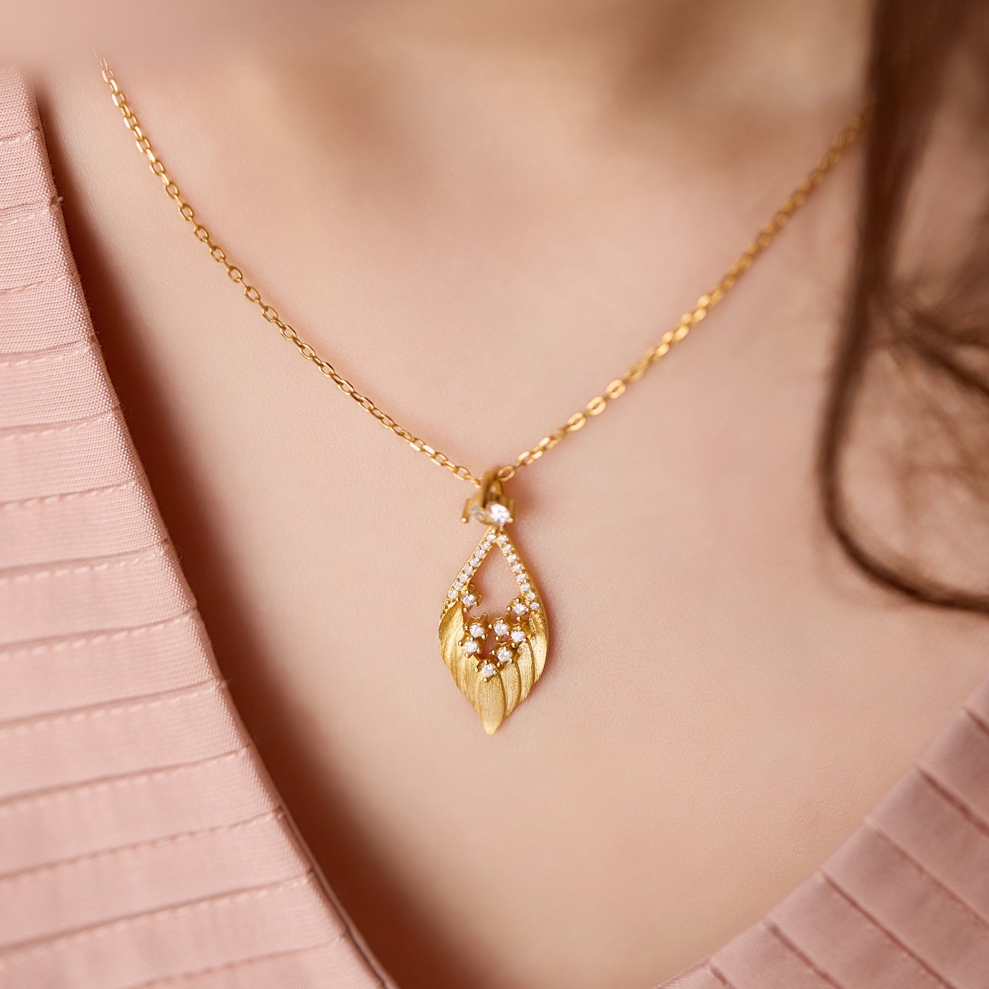 Gold necklace with a leaf-shaped pendant worn by a person against a soft pink background