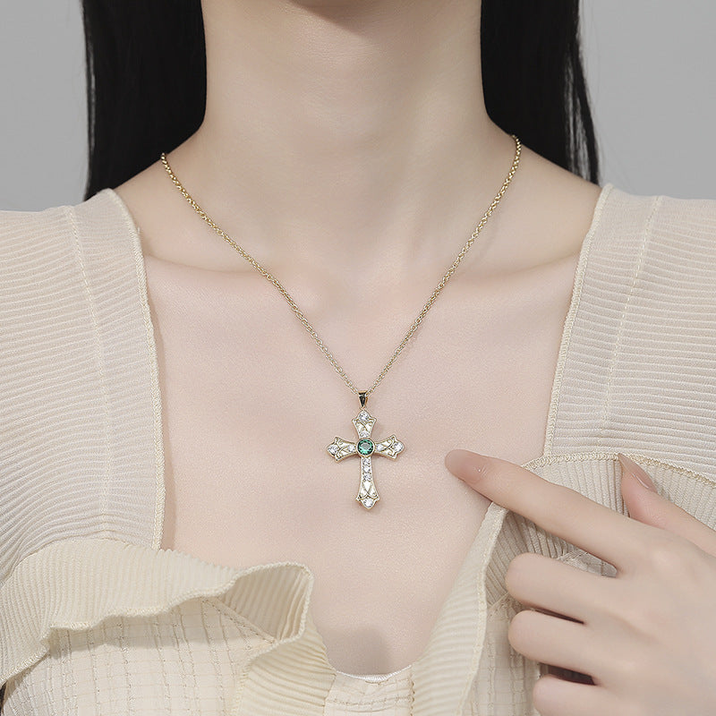 Person wearing a gold cross necklace with a green gemstone, pointing at it.
