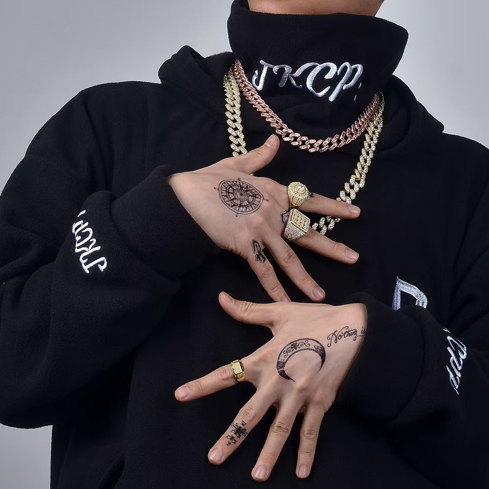 Person wearing a black hoodie with visible branding, displaying tattoos on hands and wearing gold jewelry.