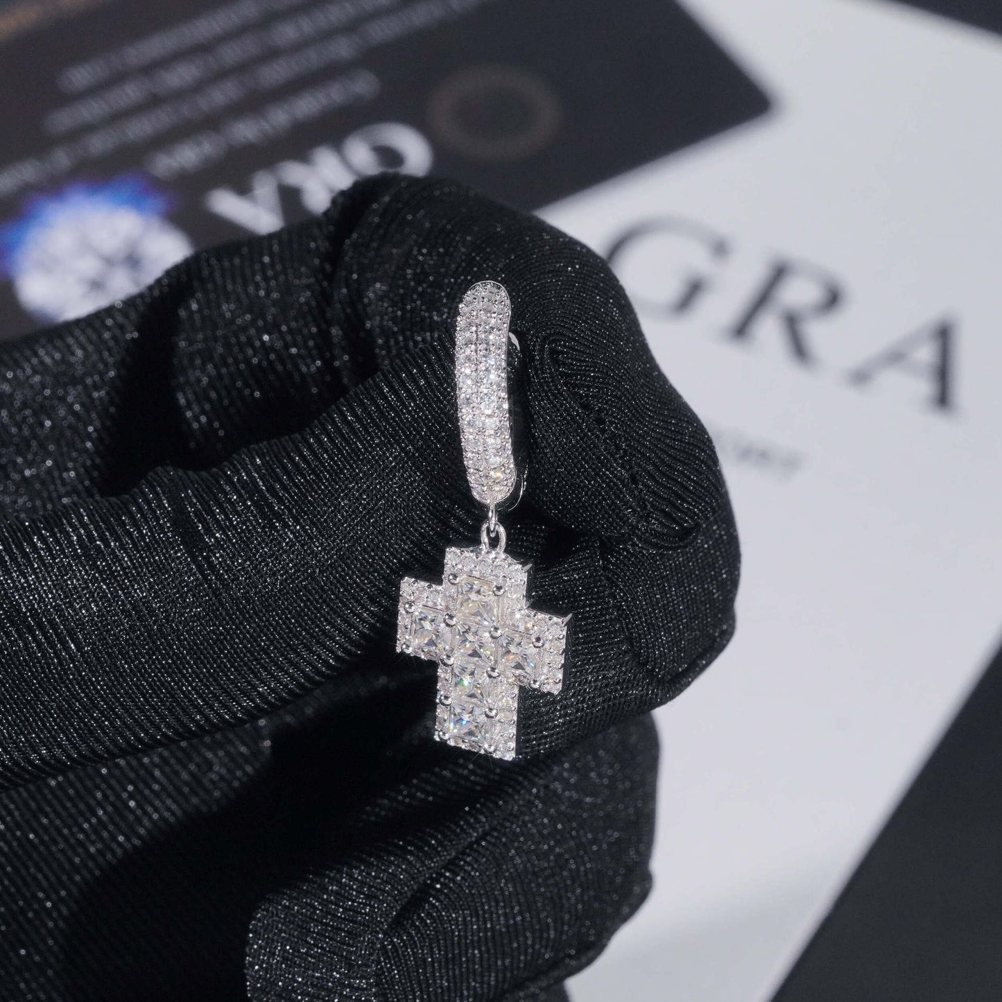 Silver cross-shaped earring held by a gloved hand on a black and white background