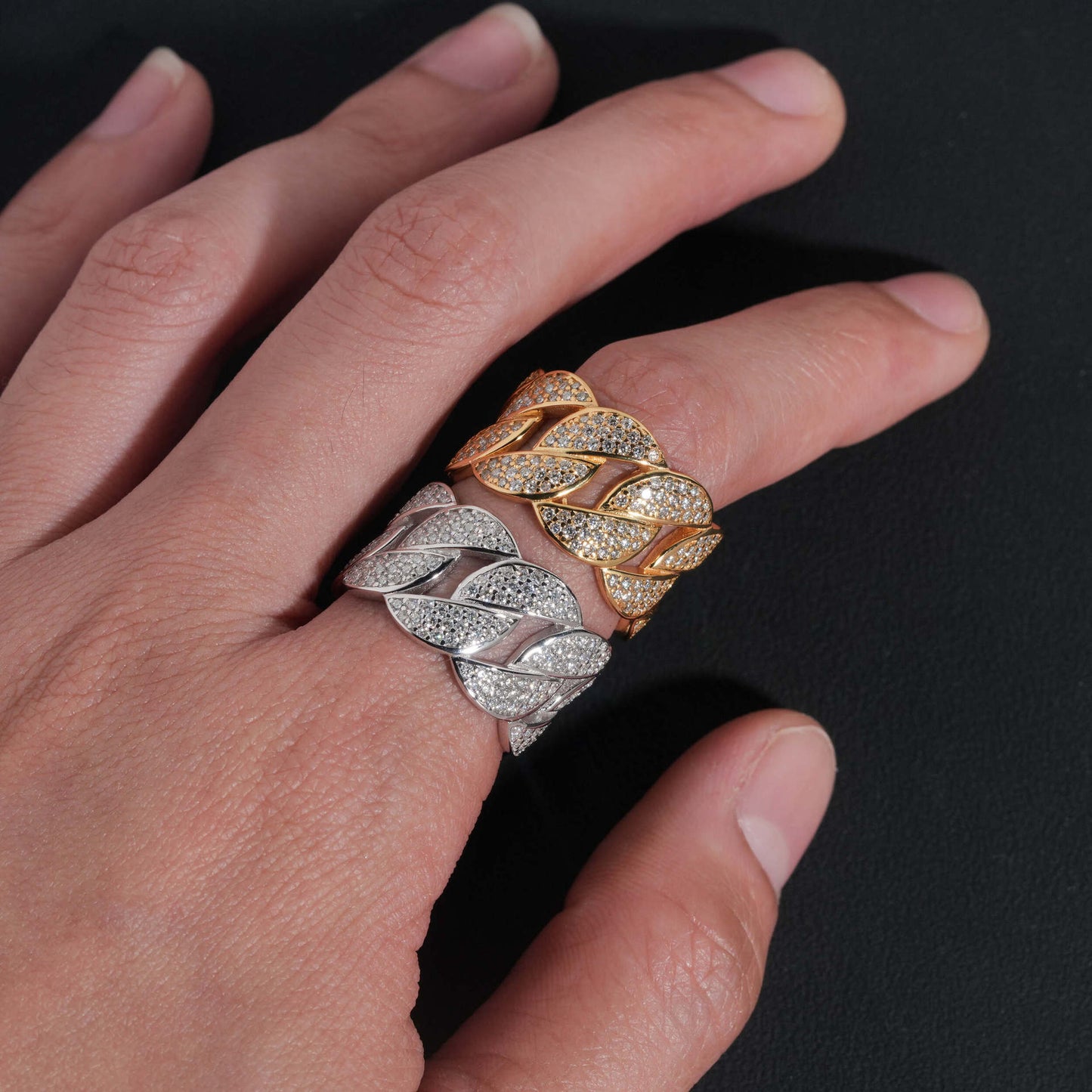 Hand wearing two braided rings, one gold and one silver, on a dark background