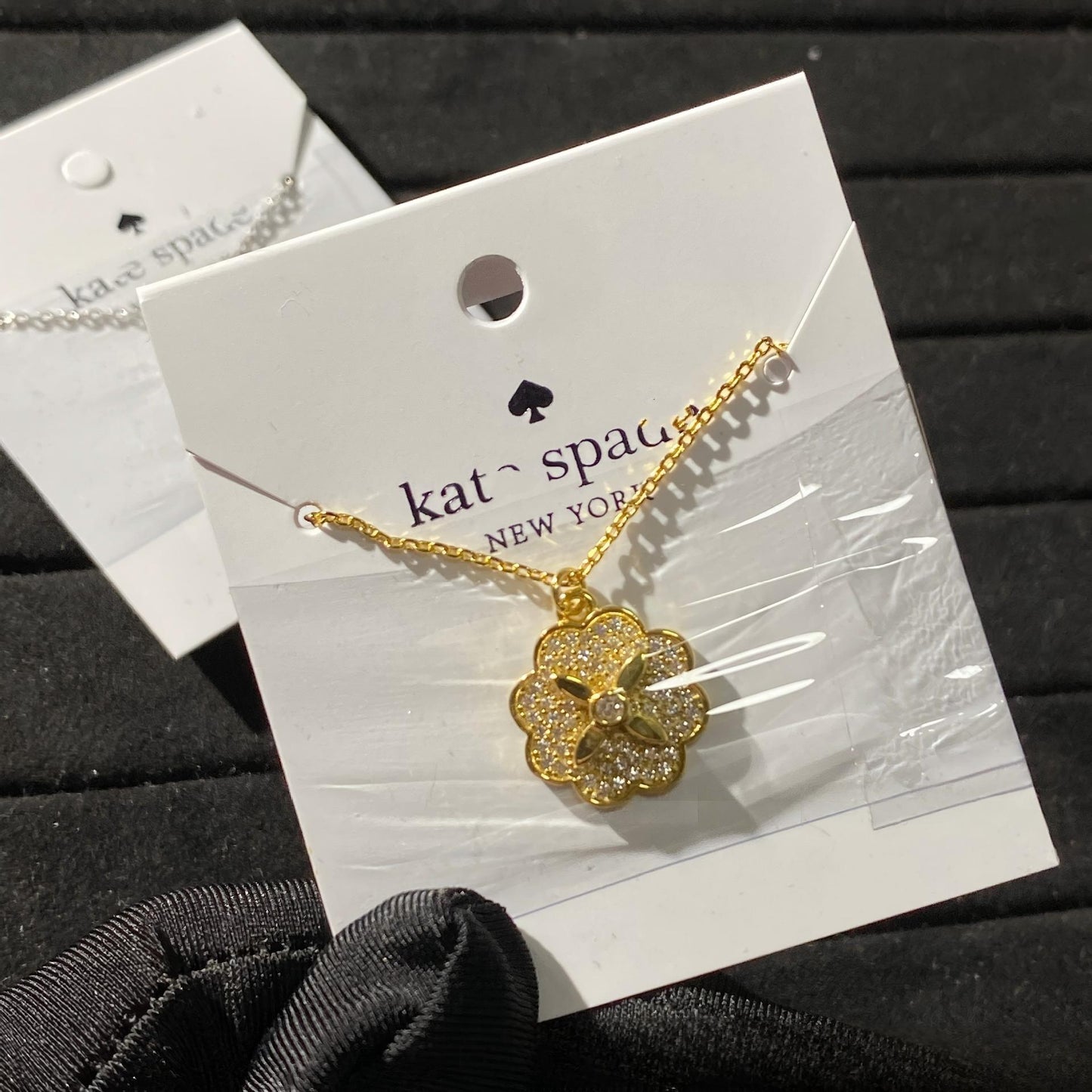 Clean white or soft pastel background — the Kate Spade Heritage Bloom mini pendant displayed on a bust form. The floral enamel pendant catches soft light, showing delicate petal details and the signature Kate Spade aesthetic.