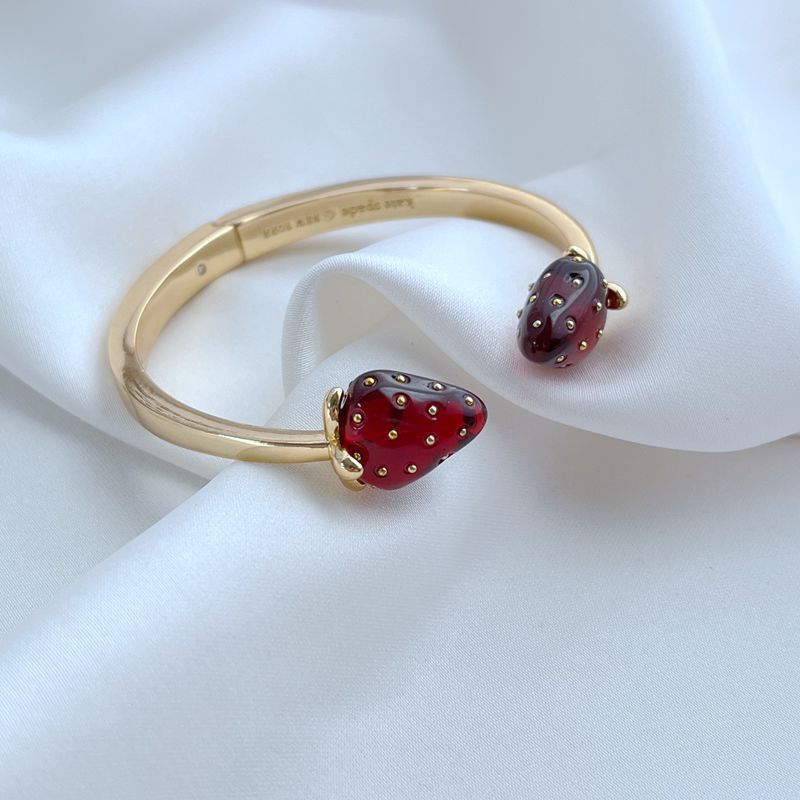 Bracelet on a light, natural background (white wood or pale pink), highlighting the strawberry enamel details. Alt: “Kate Spade Strawberry Gold Bracelet – Dainty Fruit Charm Jewelry.”
