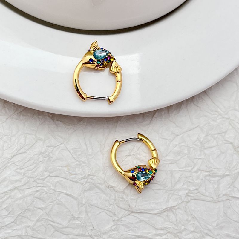 Earrings on a soft aquatic blue or seafoam green background, showcasing the gold huggie hoop with dangling enamel fish charm. Alt: “Kate Spade What A Catch Fish Huggie Earrings NEW – Gold Fish Hoop Jewelry.”