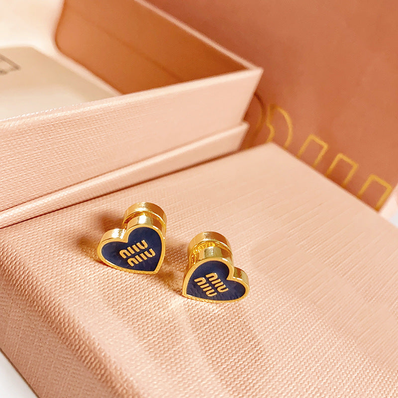 A sharp, close-up shot on a crisp white or light grey background, clearly displaying the heart logo's black enamel and gold metal contrast. Alt text: "Miu Miu Black and Gold Heart Logo Stud Earrings - Iconic Designer Studs."