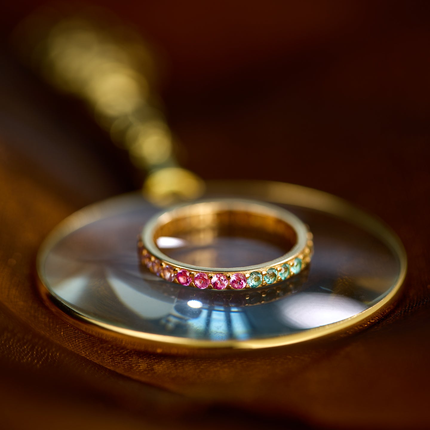 Gold ring with colorful gemstones under a magnifying glass on a wooden surface