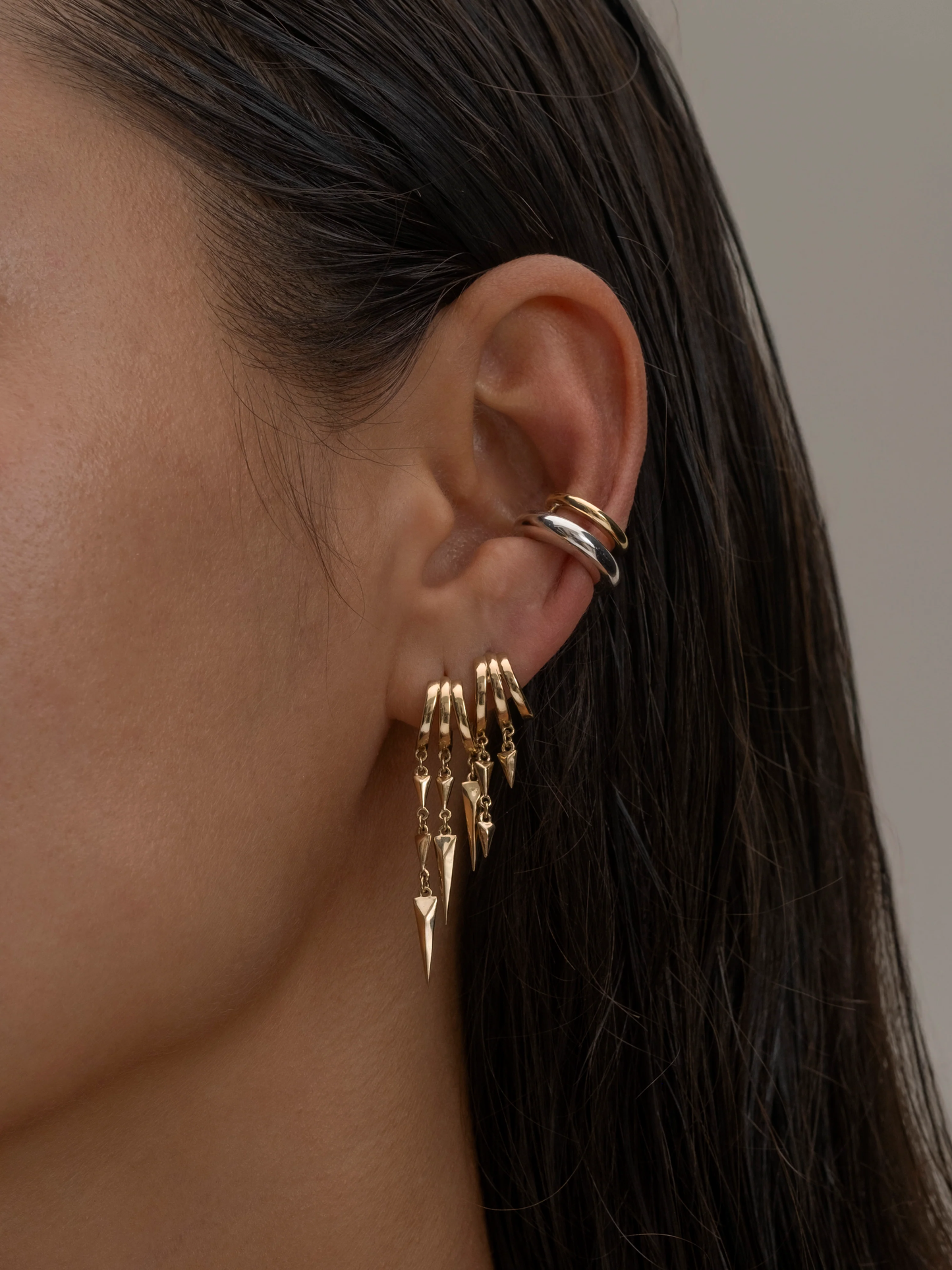 Close-up of an ear with gold S925 Sterling Silver Lynx Stacked Triple Hoop Huggie Illusion Earringsand a ring, against a neutral background