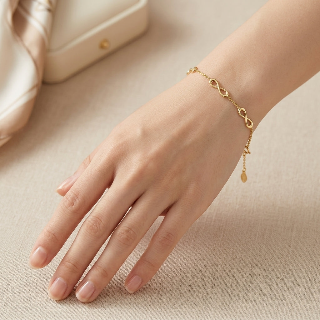 A delicate gold chain bracelet with an infinity loop design and a small dangling teardrop charm, shown worn on a woman's wrist against a soft beige background.