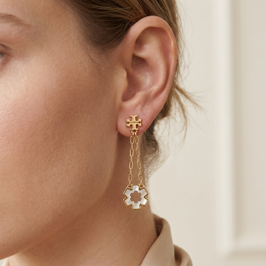 A close-up of a model's ear wearing a gold dangle earring with a small cross stud, chain link, and a white enamel flower charm.