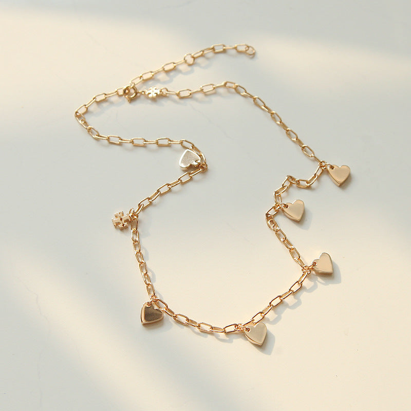 A delicate gold chain anklet featuring five small heart charms of varying sizes, displayed on a light cream surface with soft, natural shadows.