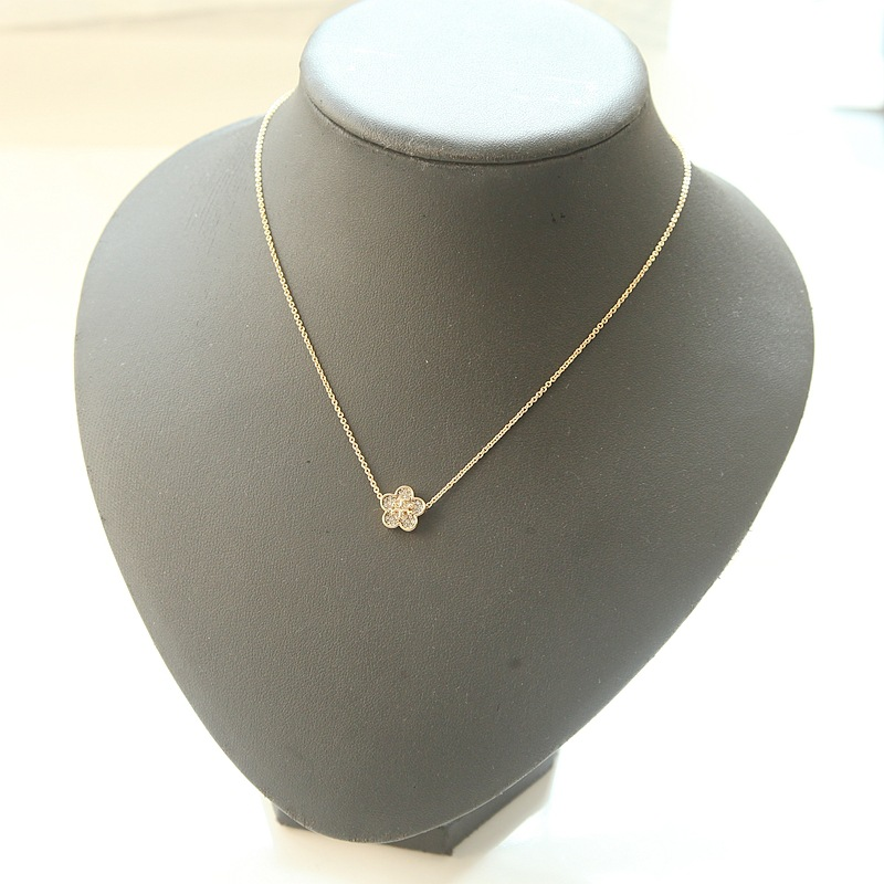 A delicate gold chain necklace with a small flower-shaped pendant, displayed on a matte black jewelry bust against a bright white background.
