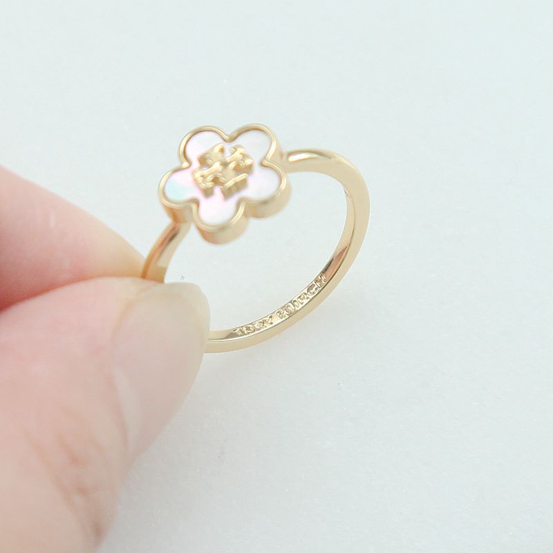 A gold ring with a small flower-shaped charm featuring iridescent white inlay and a gold star in the center, held by a person's fingers against a plain light background.
