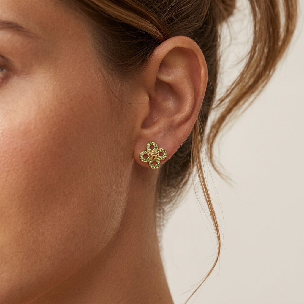 A close-up of a woman's ear wearing a small gold four-petal flower stud earring with tiny red and green gemstone accents, against a light neutral background.