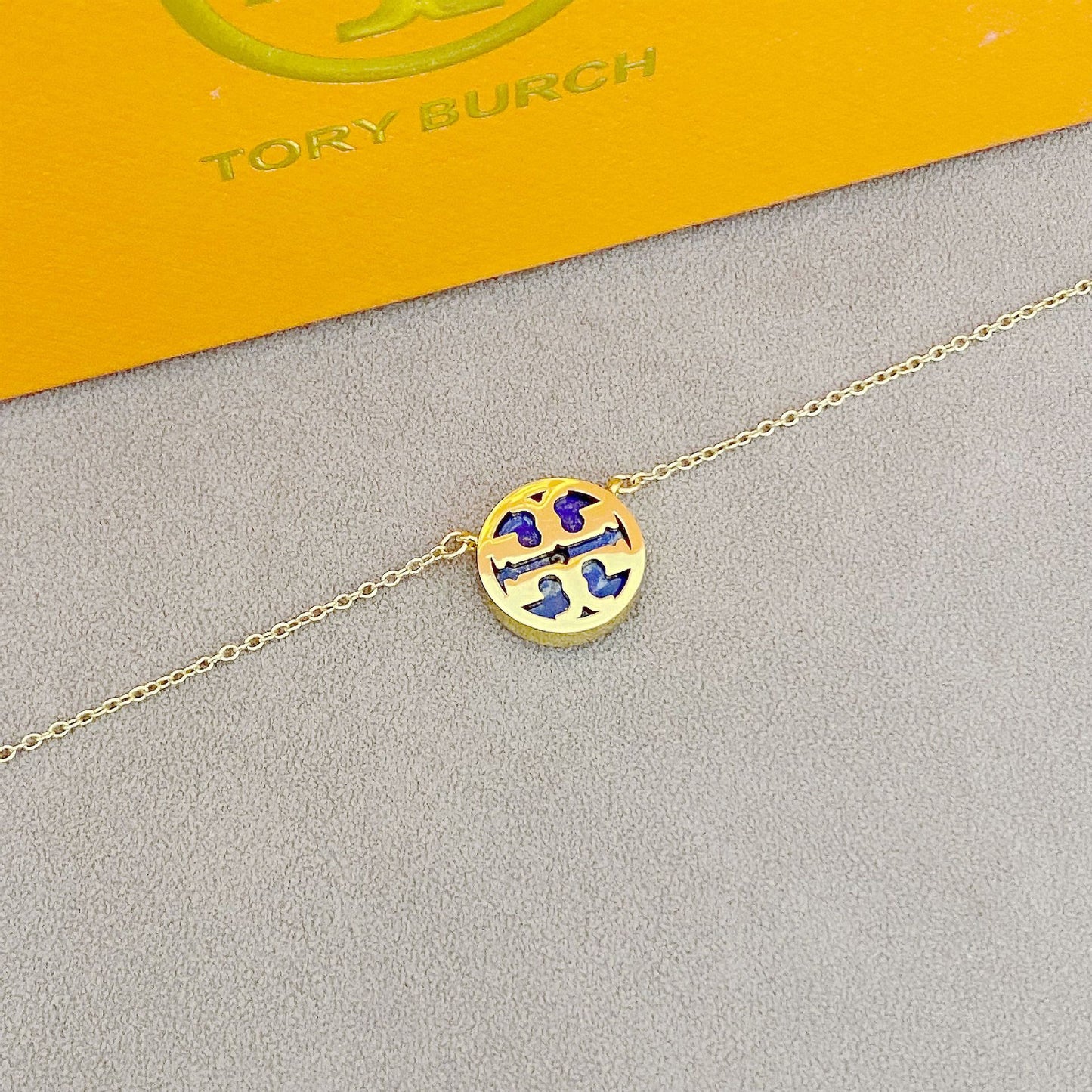 Gold necklace with a round pendant on a gray surface, with 'Tory Burch' branding in the background.