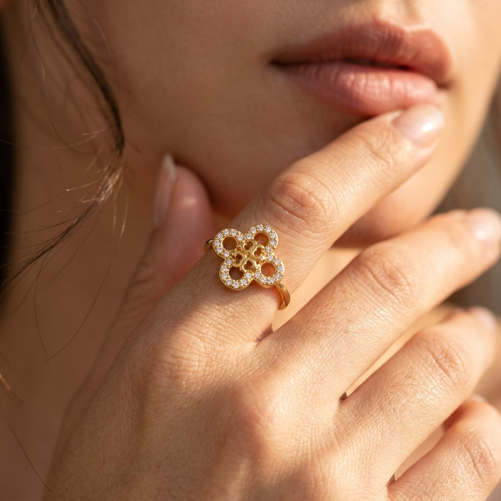 A close-up lifestyle shot of a woman's hand wearing a gold ring with a flower-shaped pavé setting. The warm sunlight highlights the delicate metal and clear stones, with the woman's lips and face softly blurred in the background.