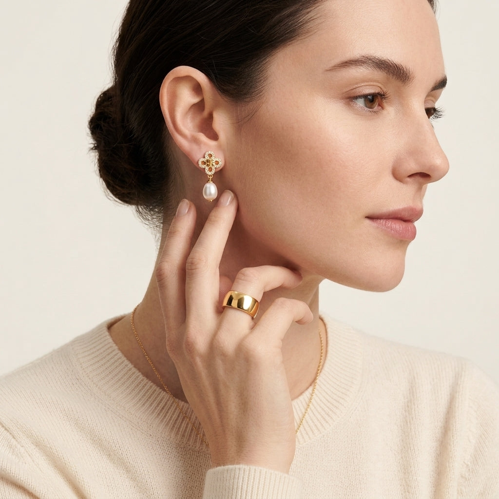 A close-up of a woman wearing a delicate gold earring with a white teardrop pendant and a gold ring, set against a soft beige background.