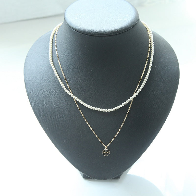 A double-layered necklace set displayed on a black jewelry bust. The top layer features a delicate strand of white pearls, while the bottom layer is a fine gold chain with a small gold clover-shaped pendant.