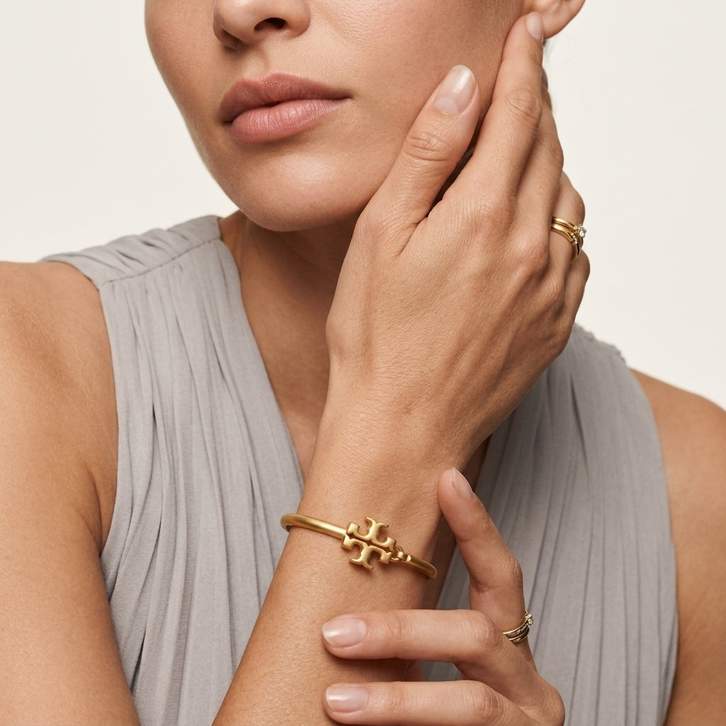 A close-up lifestyle shot of a model wearing a gold bangle bracelet with a bow-like central motif on her wrist, along with gold rings on her fingers, against a neutral light beige background.
