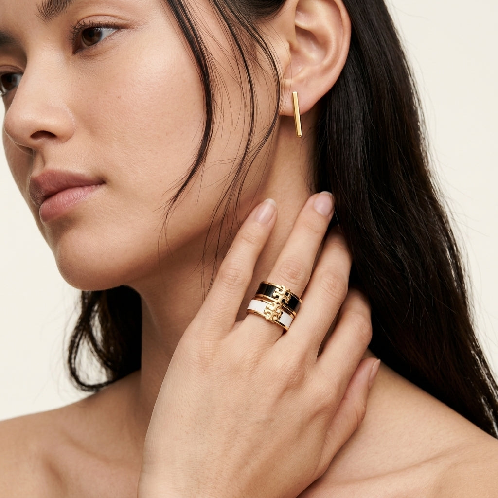 A close-up lifestyle shot of a model wearing a stack of three gold rings on her finger, with a matching gold bar earring in her ear. The rings feature textured and patterned gold bands.