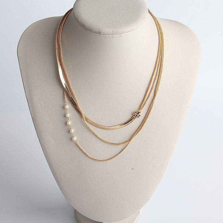 A multi-layered gold necklace displayed on a cream-colored mannequin bust. The necklace features three layers: a fine chain, a slightly thicker snake chain with a small gold flower charm, and a delicate chain adorned with a row of white pearls.