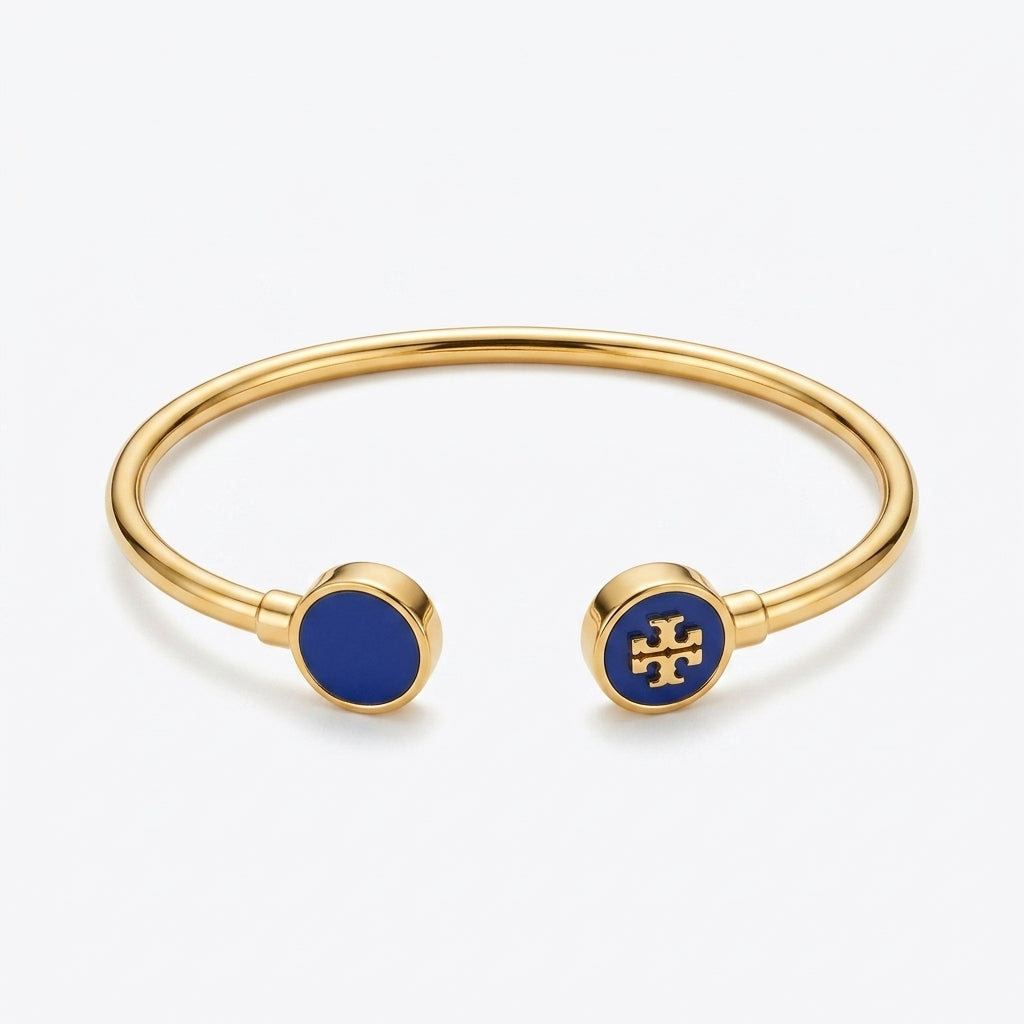 A gold-tone open cuff bangle bracelet with circular blue enamel end caps, one featuring a gold cross logo and the other a solid blue center, on a light gray background.