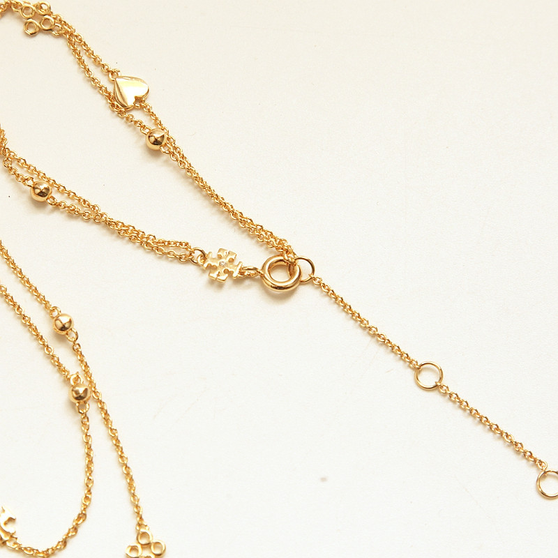 A close-up of a delicate gold chain necklace with small bead details and a tiny heart charm, laid out on a plain off-white background.