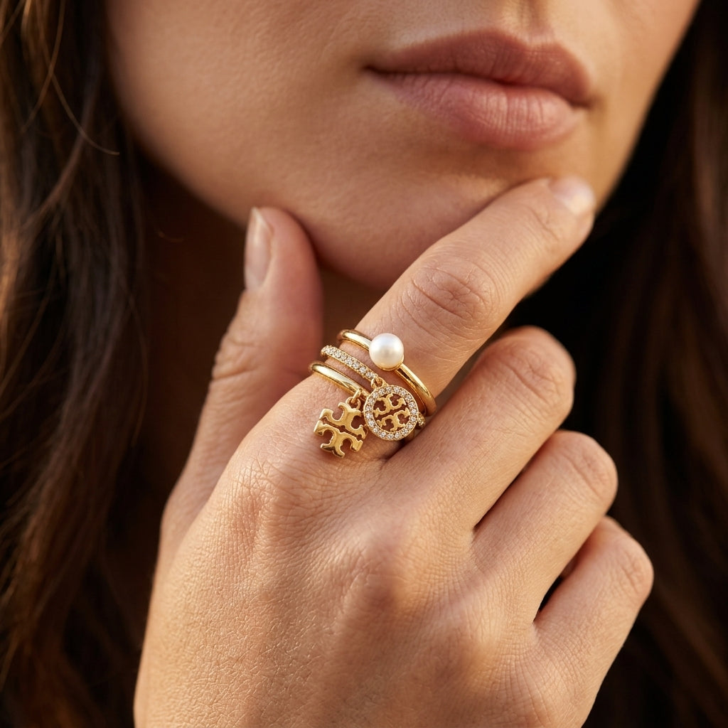 A close-up of a woman's hand resting near her chin, wearing a stack of gold rings. The rings include a gold cross charm, a gold monogram charm, a small clear crystal cluster, and a white pearl.