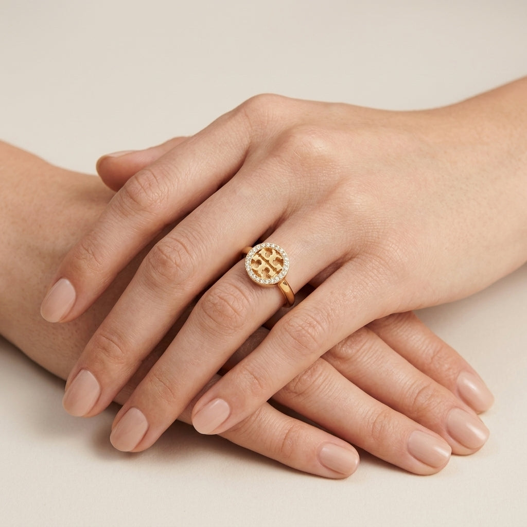 A close-up of a person's hands resting on a light beige surface, wearing a gold ring with a circular geometric cutout design and a halo of small clear stones.