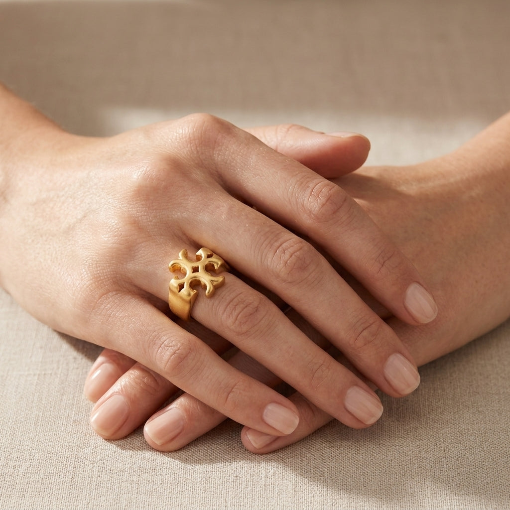 A close-up of crossed hands wearing a textured gold ring with a floral or snowflake-like openwork design, set against a soft beige fabric background.