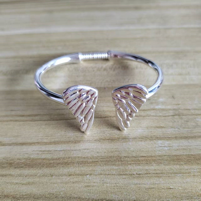 A silver open cuff bracelet with two textured wing-shaped charms, displayed on a light wood grain surface.