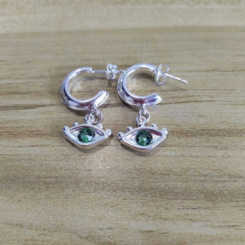 A pair of silver hoop earrings with dangling eye charms featuring green gemstone centers, displayed on a light brown wooden surface.
