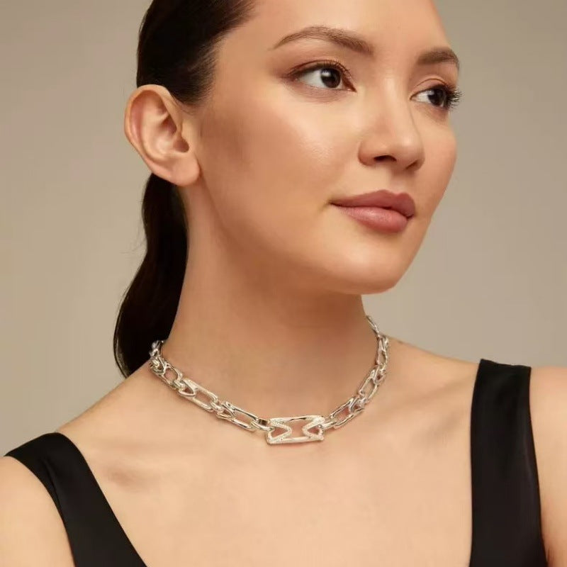 A close-up of a woman wearing a silver safety-pin chain necklace with a polished finish, paired with a black top against a neutral beige background.