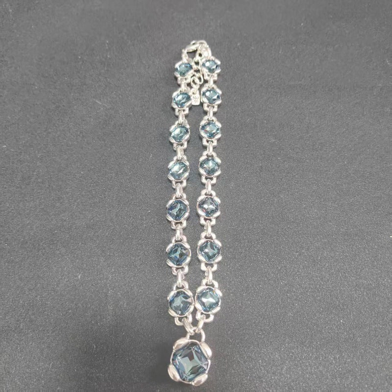 A silver-toned double-strand chain bracelet featuring small light blue faceted gemstones and a larger matching blue pendant at the bottom, displayed on a dark gray textured background.