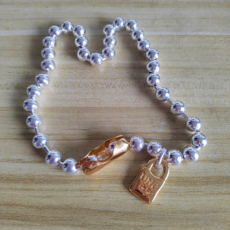 A silver beaded bracelet with a gold-toned padlock charm engraved with 'UNG KEE', displayed on a light wood background.