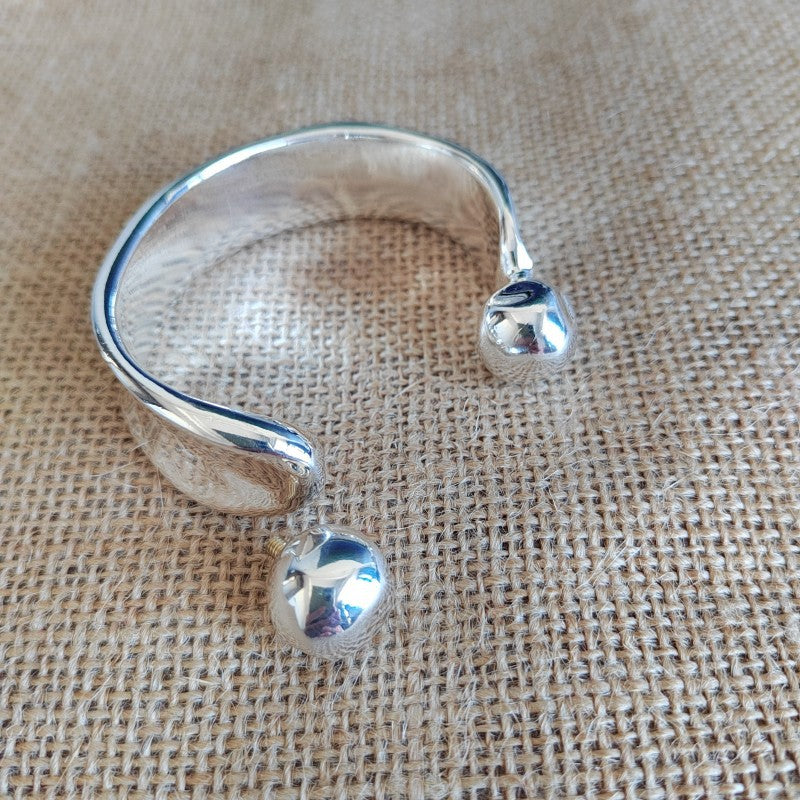 A polished silver-tone open cuff bracelet with two small spherical bead details, displayed on a textured beige burlap fabric background.