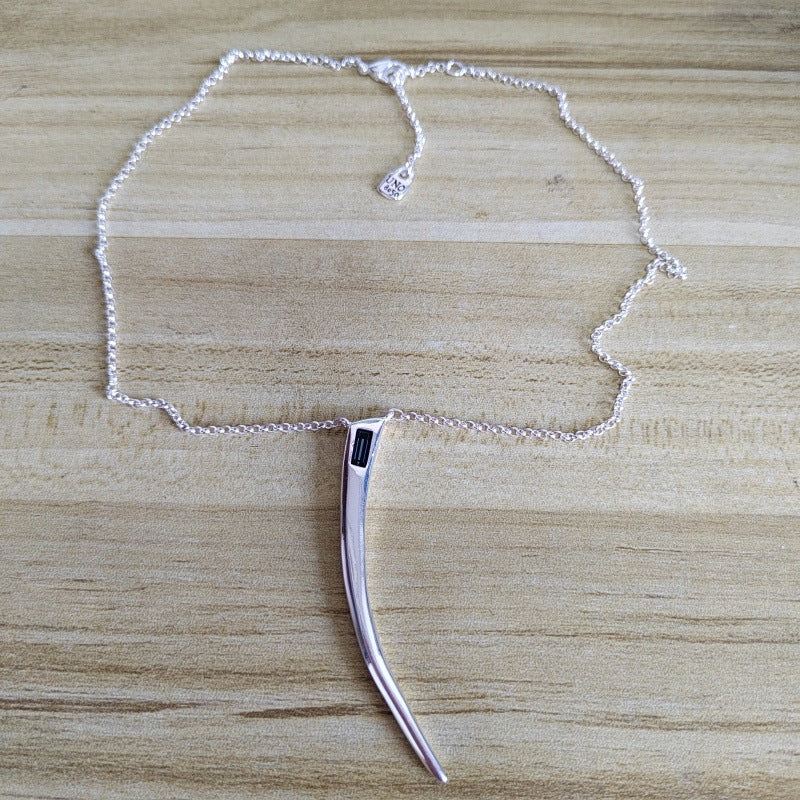 A silver-toned necklace with a curved, knife-like pendant featuring a small black rectangular accent, displayed on a light wood grain surface.