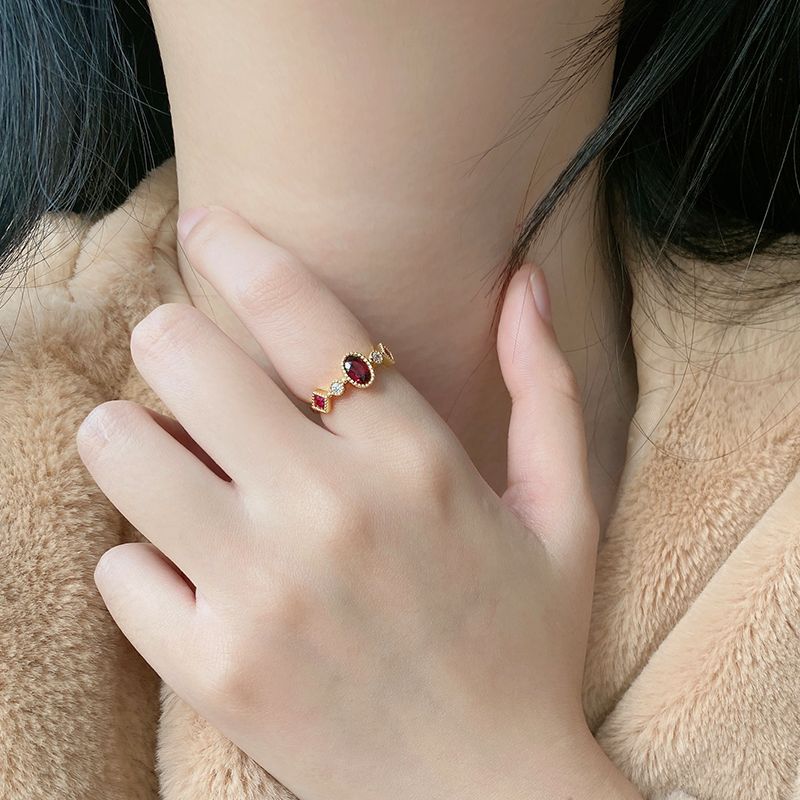 Close-up of a hand wearing a gold ring with a red gemstone, held against a soft beige fabric background.