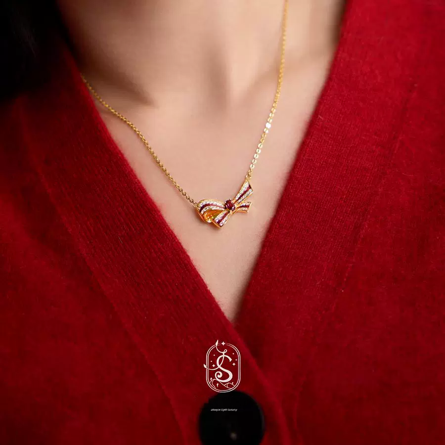 Gold necklace with a heart-shaped pendant worn by a person in a red garment, with a brand logo at the bottom.