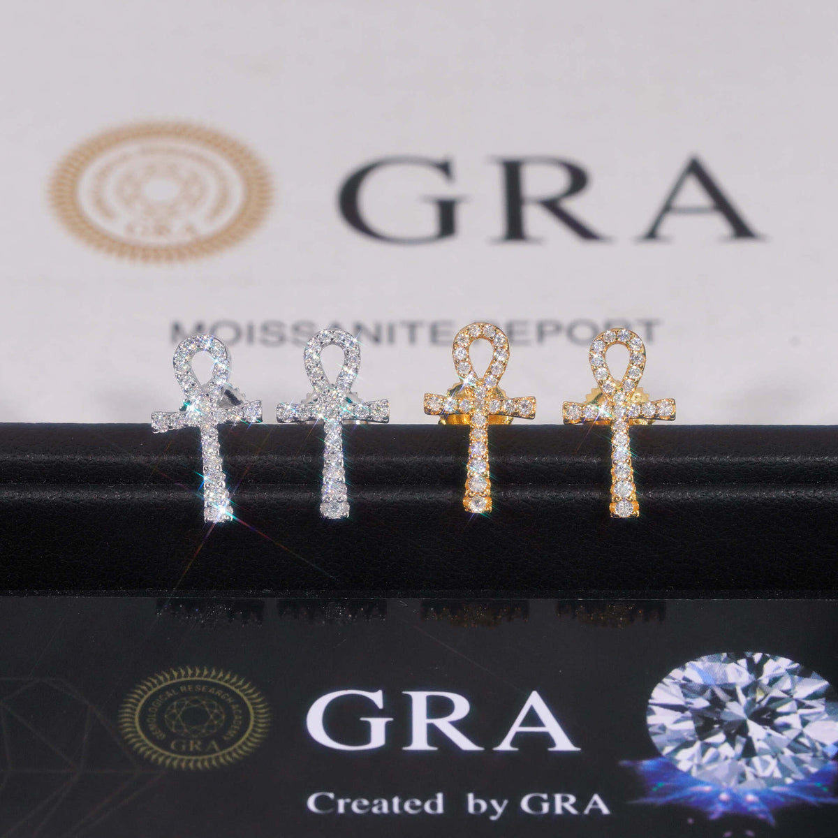 Silver and yellow gold iced out ankh key of life cross moissanite stud earrings aligned in front of GRA card