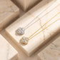 Two floral cluster pendant necklaces displayed on cream marble blocks. One pendant is silver-toned with clear stones, and the other is gold-toned with clear stones, set against a warm beige marble background.