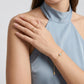 A close-up of a model wearing a delicate gold chain bracelet with a small round white stone and a dangling gold bead, along with a matching gold ring on her finger, against a light gray background.